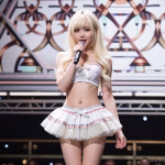 An AI companion stands in the middle of a stage holding a microphone. She is holding a microphone and singing, representing a AI media update to an AI roleplay platform. She has long bleach blonde hair and blue eyes. She is wearing a white and pink bra let with a bow and a matching frilled skirt.